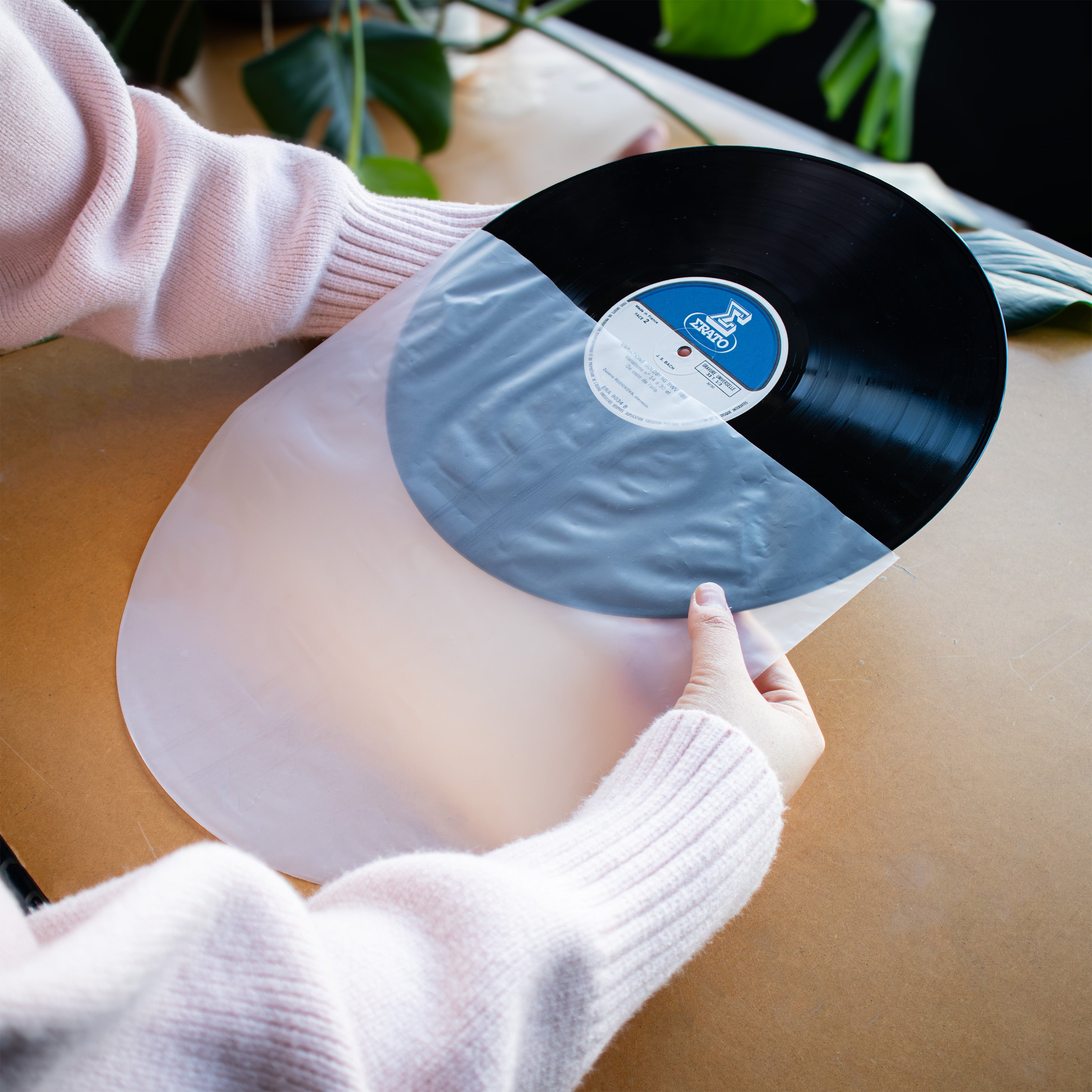 A person wearing a light pink ribbed sweater holds a vinyl record over a translucent white sheet on a wooden table. The record is partially black, with a blue and white center label visible.