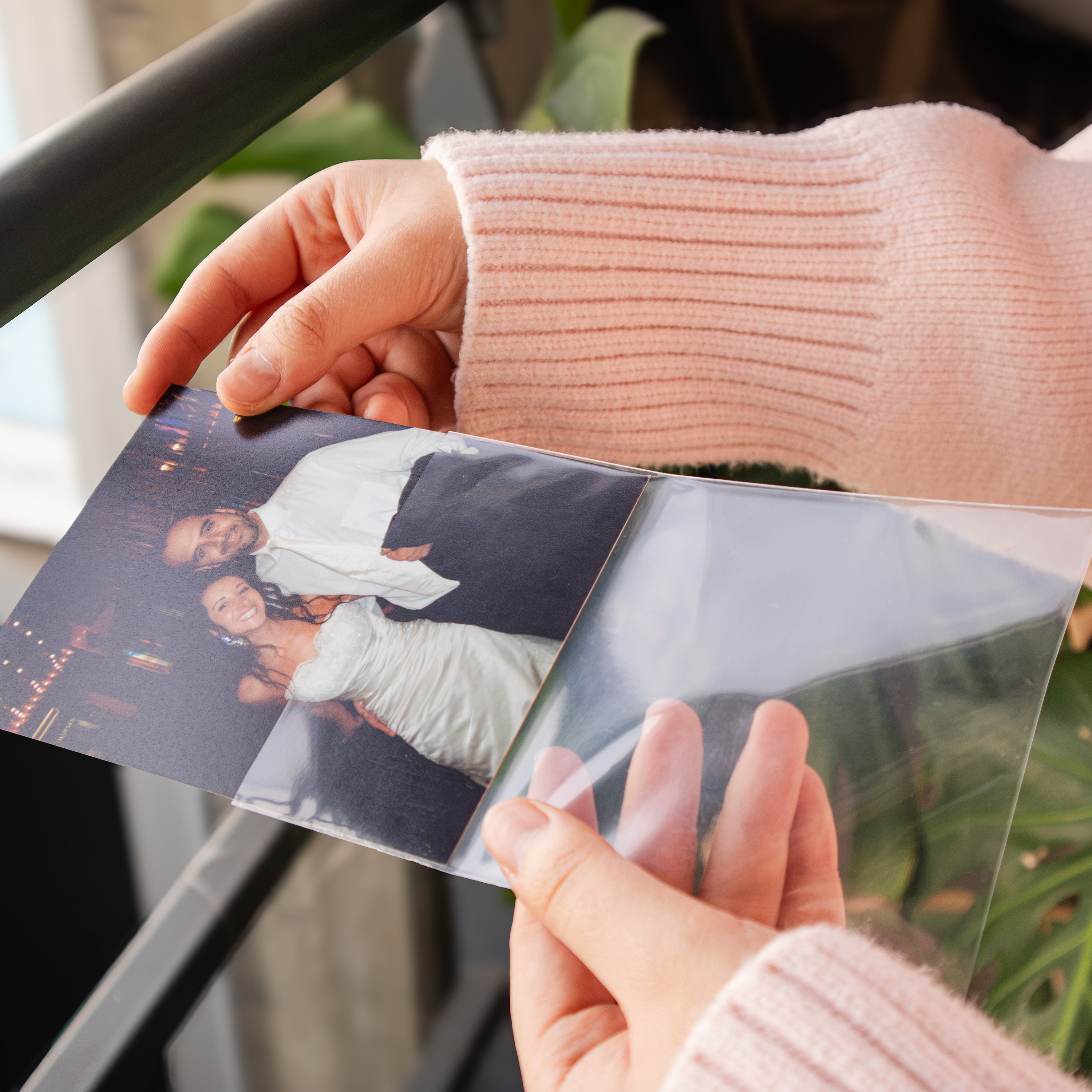 A close-up of hands holding a clear plastic photo album or binder, showing printed photographs inside. The person is wearing a light pink ribbed sweater, and the background includes a dark railing and green plants.
