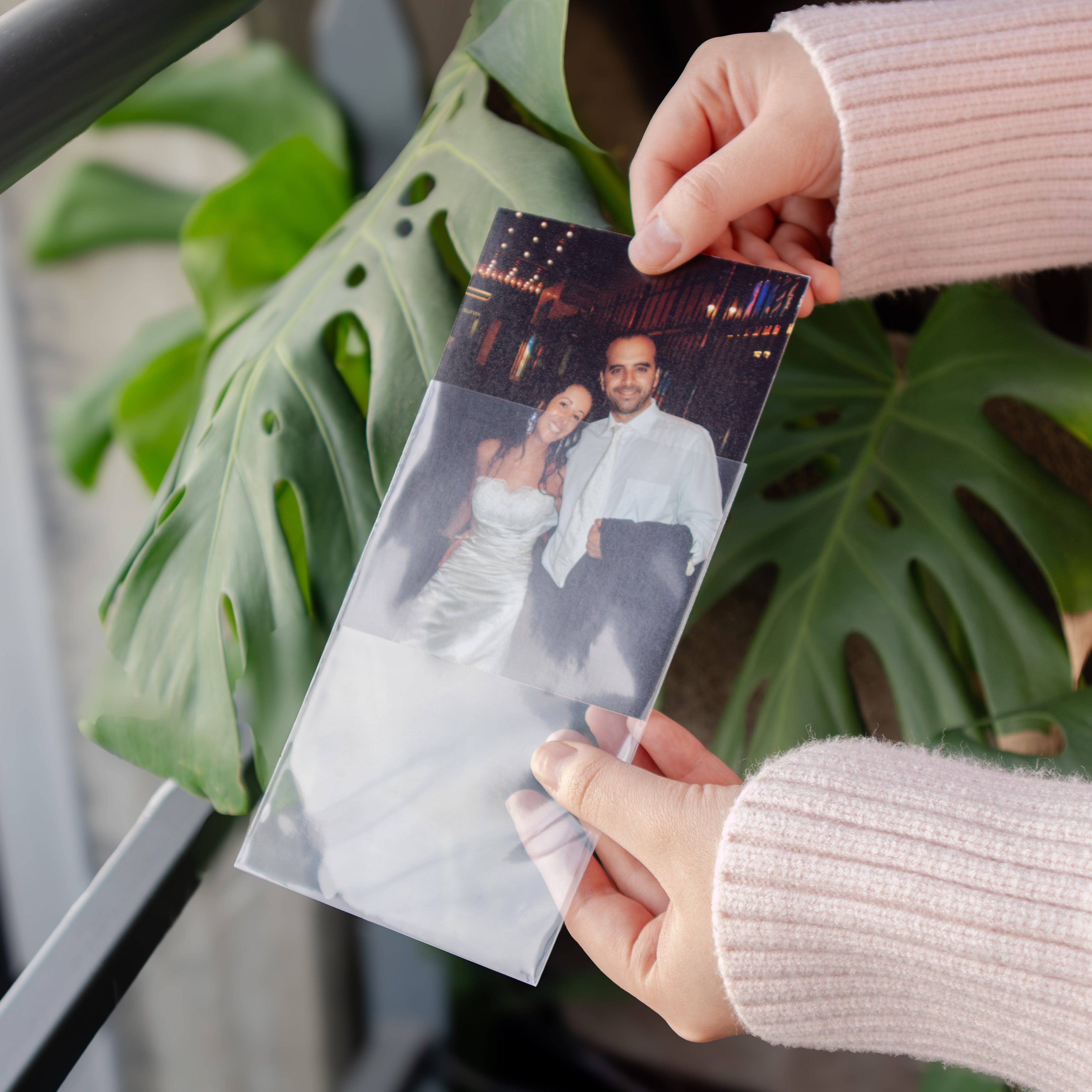 A person holding a long, narrow printed photo strip or card with a faded photograph of a couple, photographed in front of large green monstera leaves. The person is wearing a light pink ribbed sweater, and the product itself appears to be the printed photo strip.