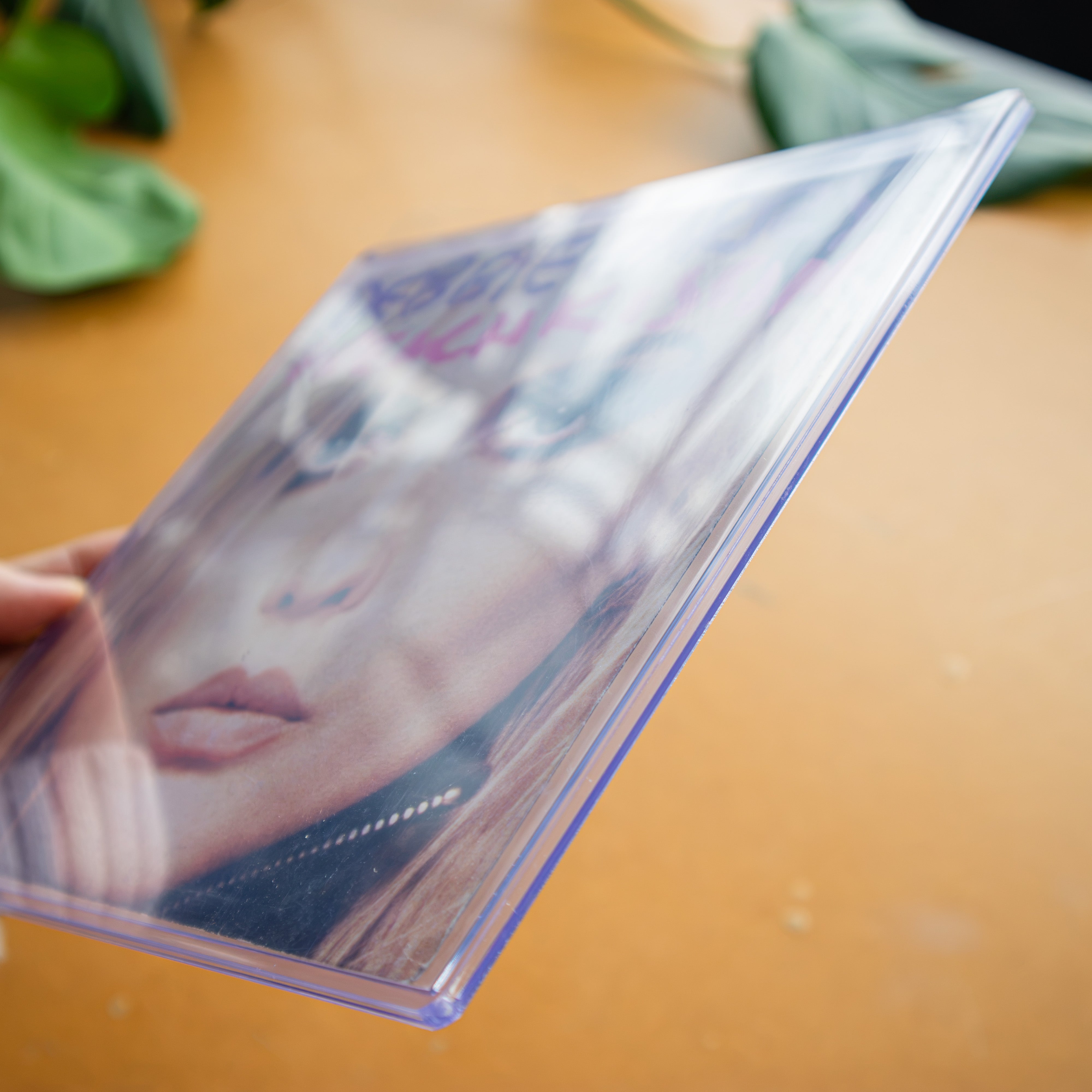 A hand holding a clear plastic photo frame with a printed portrait insert, photographed over a warm tan surface with blurred green leaves in the background.