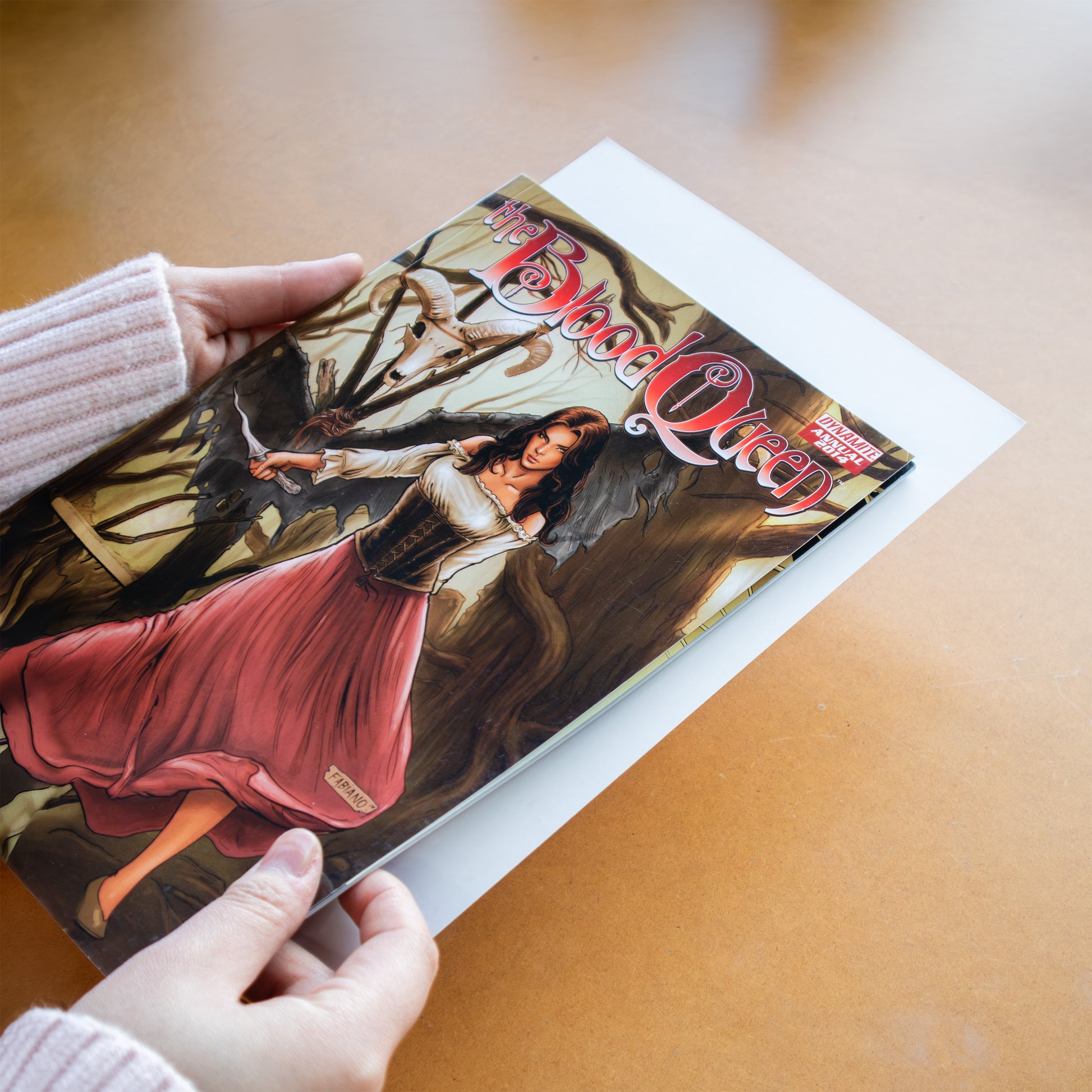 A person holds a comic book titled 'The Blood Queen' comic over a white comic board on a warm wooden surface.