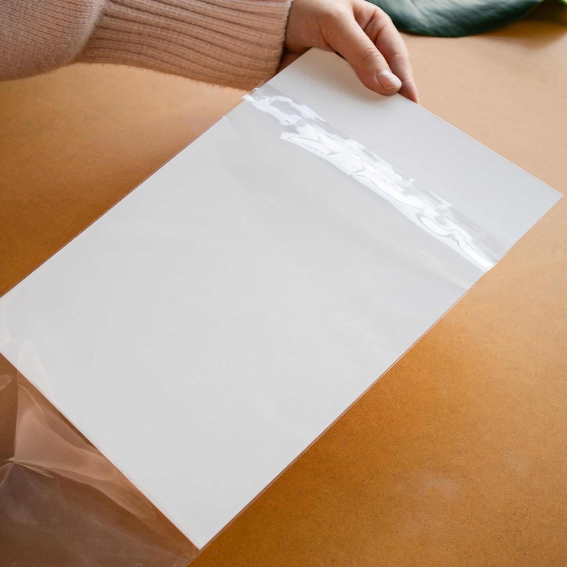 A hand holding a large, clear rectangular sheet of transparent plastic or acrylic over a warm brown surface.