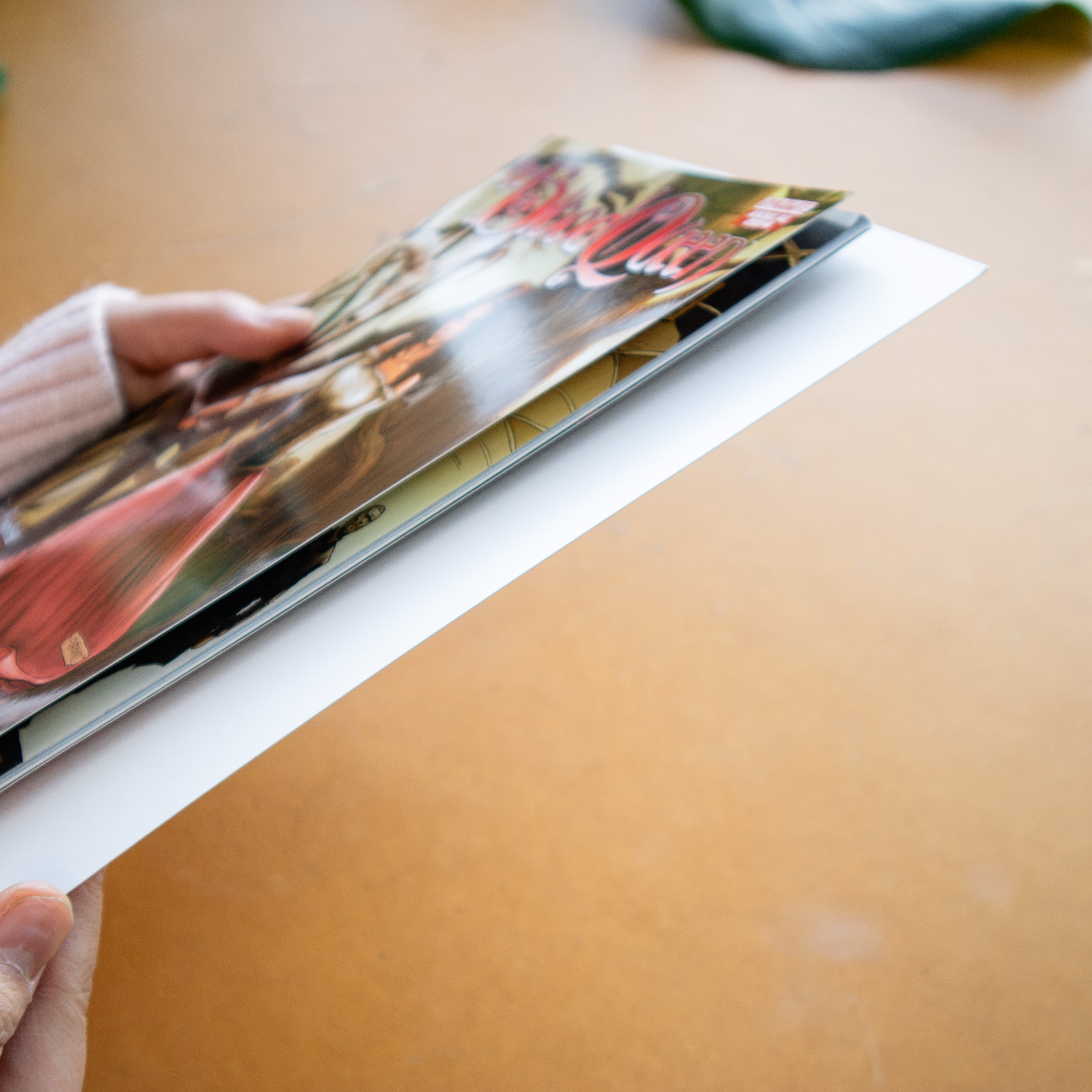 A hand holds a glossy printed comic over a white comic board on a warm tan surface.