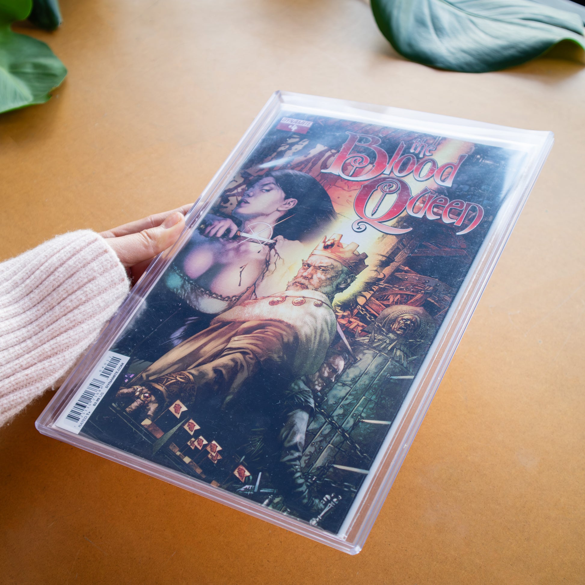 A comic book in a clear protective comic toploader being held by a hand, placed on a warm brown surface with green leaves in the background. The comic cover reads 'The Blood Queen'.
