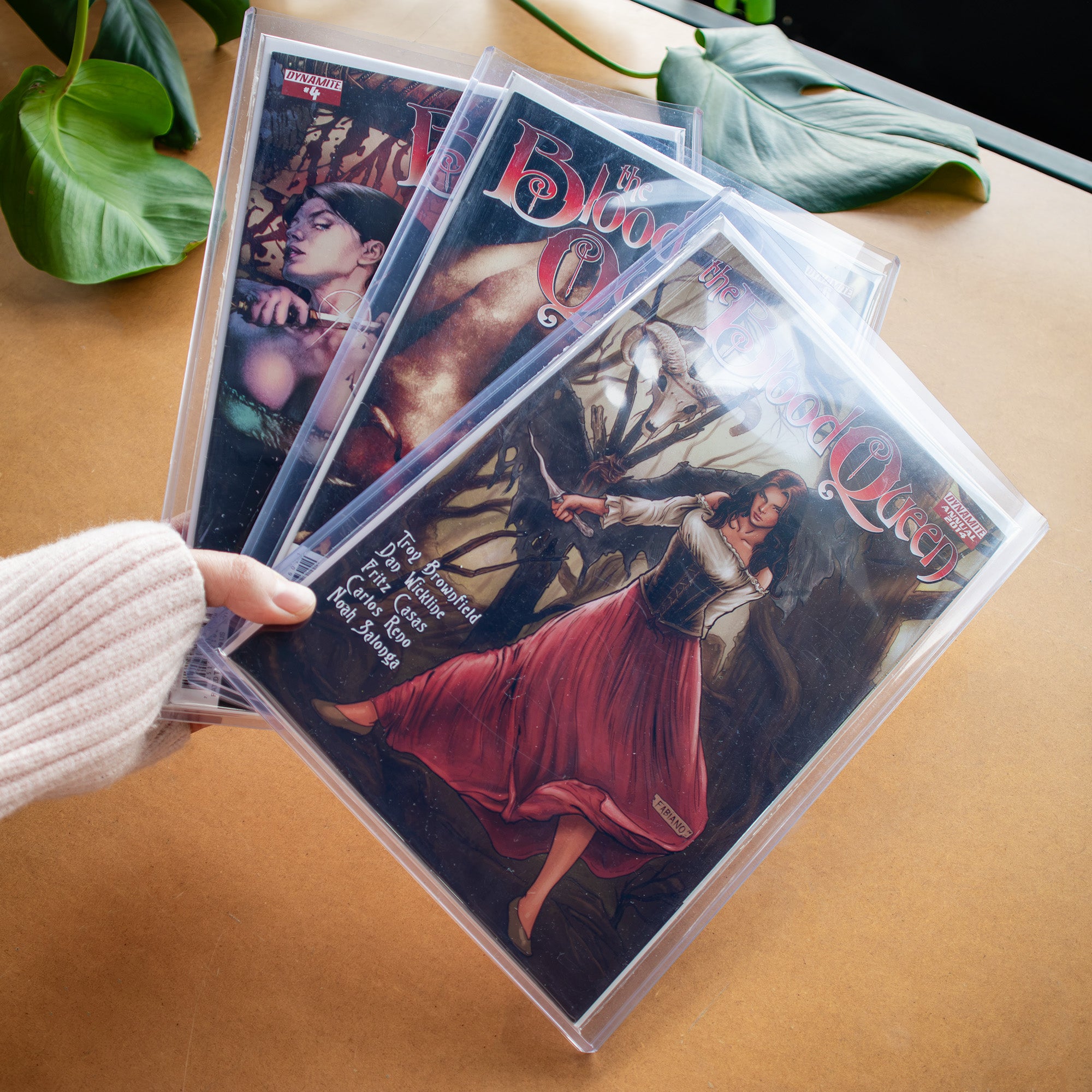 A hand wearing a cream knit sleeve holds a series of comic book in clear protective comic toploaders on a tan tabletop with green leaves in the background.