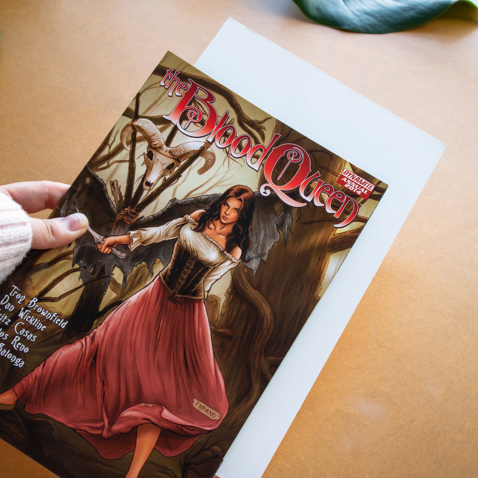 A hand holds a comic book titled 'The Blood Queen' over a white comic board on a tan surface.