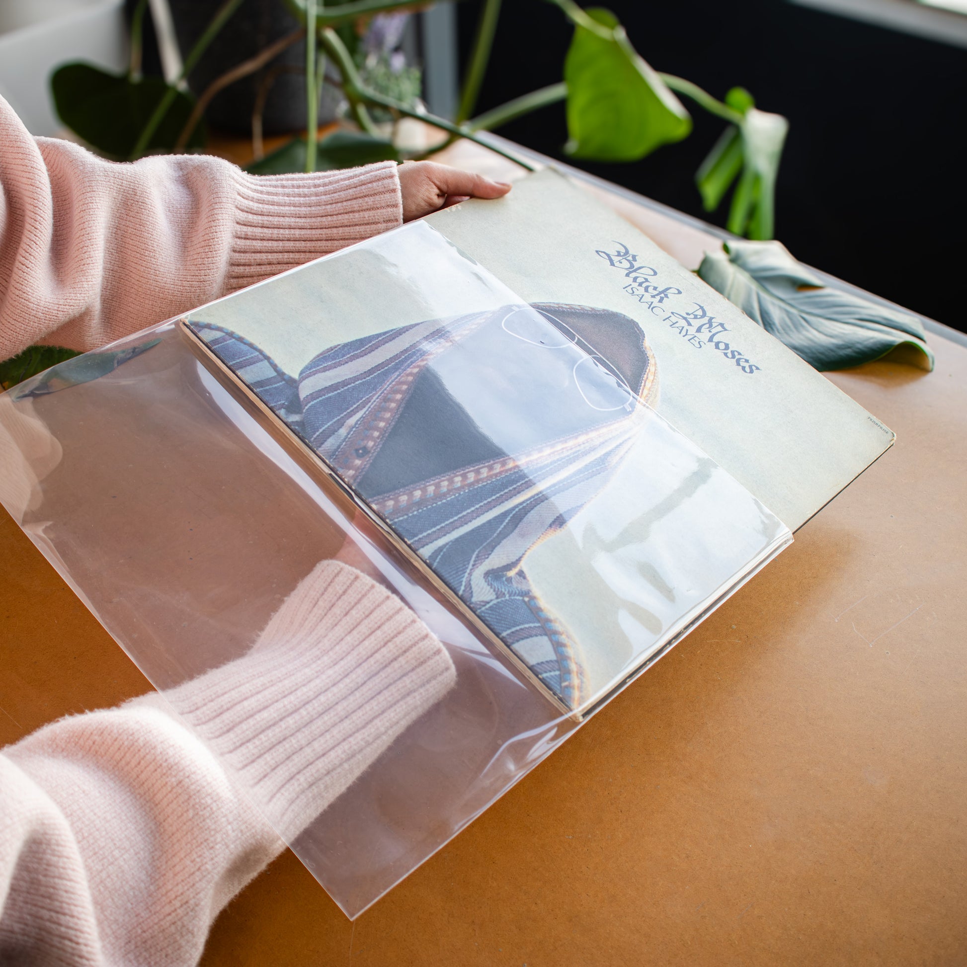 A person wearing a light pink ribbed sweater holds a clear plastic sleeve over a printed card or booklet on a warm brown tabletop, with green leaves blurred in the background.